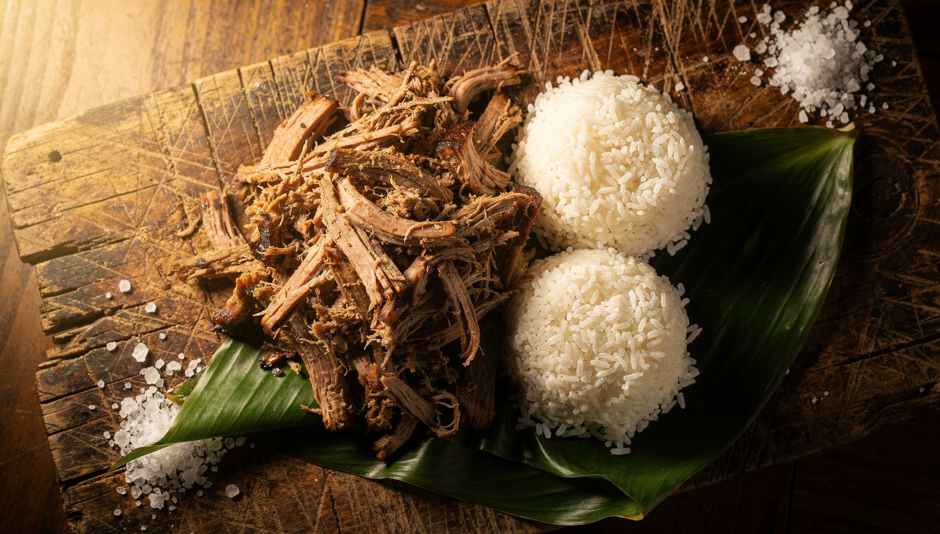 Kalua pork hawaiano desmenuzado sobre tabla de madera con arroz blanco y hojas de plátano