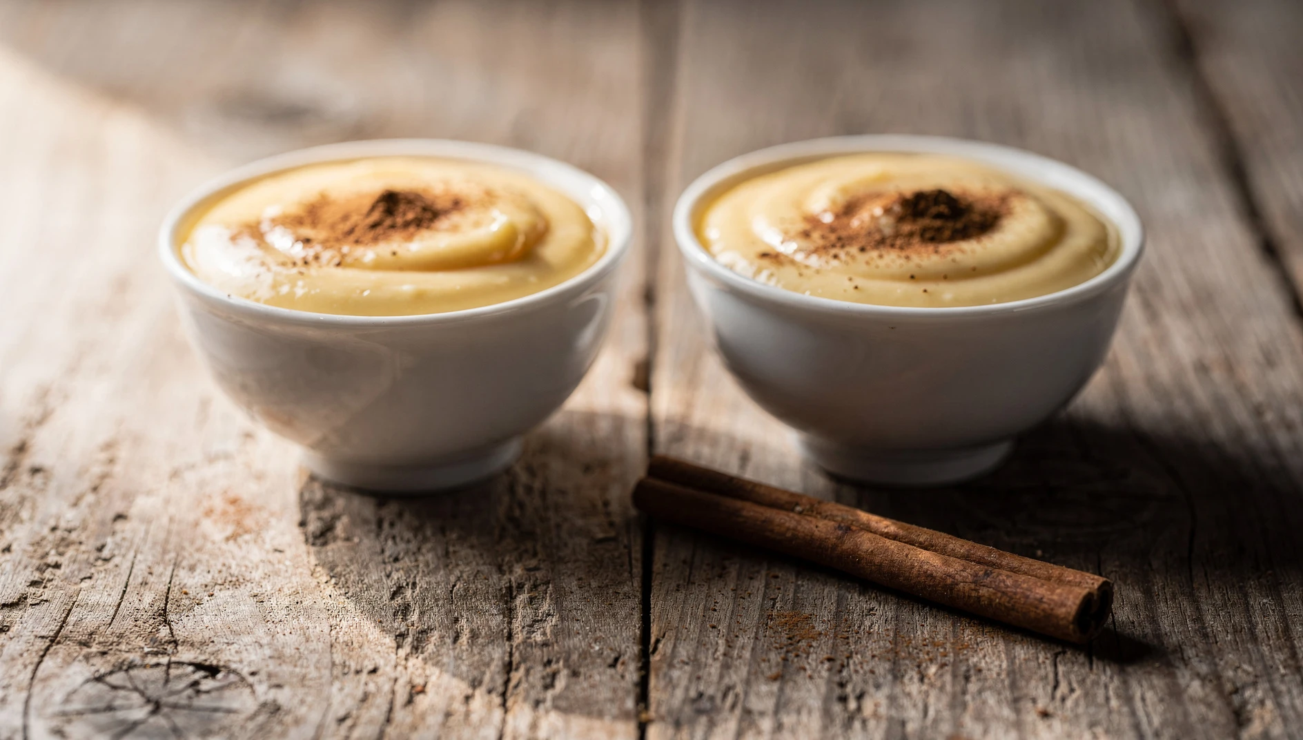 Dos cuencos de natillas veganas de vainilla espolvoreadas con canela sobre una superficie de madera