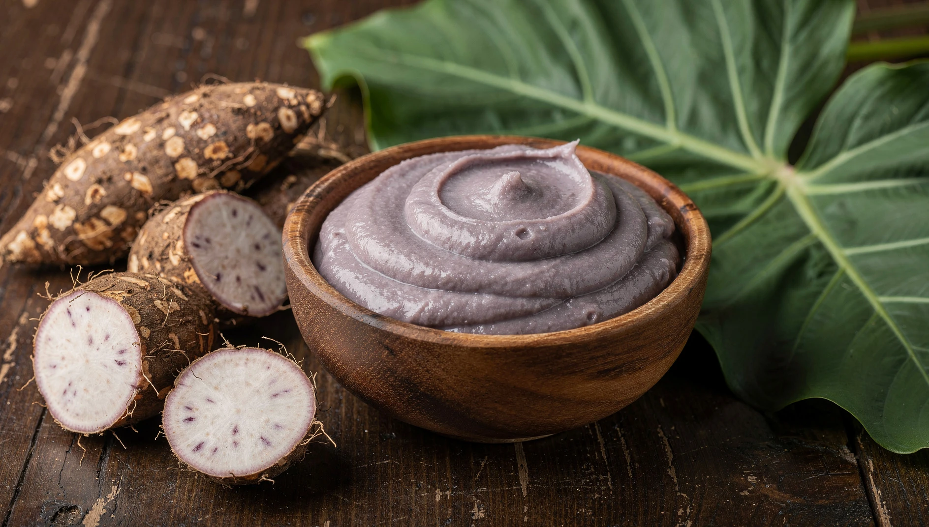 Bol de madera con poi hawaiano elaborado con raíz de taro, acompañado de raíces y hojas frescas de taro sobre mesa de madera oscura