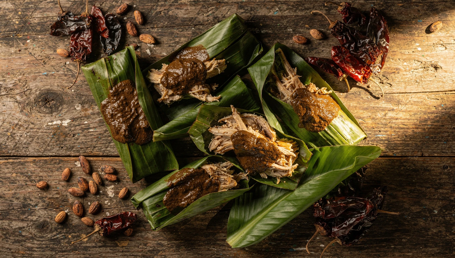 Tamales oaxaqueños de mole negro con pollo deshebrado envueltos en hoja de plátano sobre mesa de madera rústica